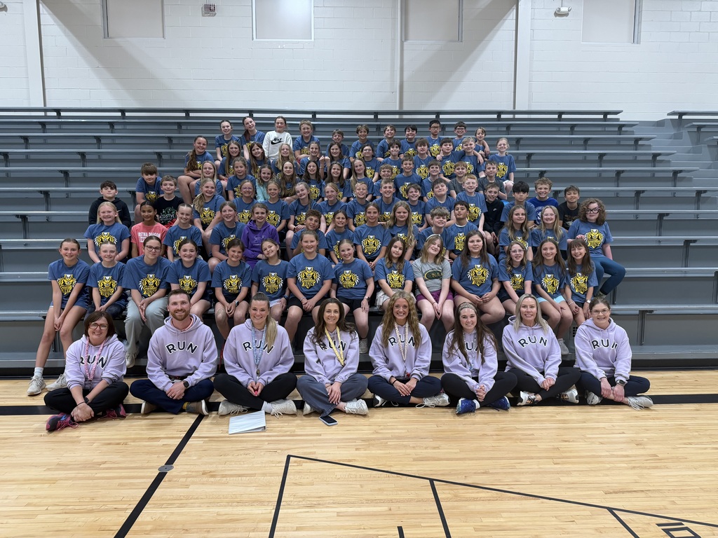 A large group portrait of children and several adults in an indoor gym, seated and standing on bleachers. Most wear matching blue t-shirts. The adults in front wear lavender hoodies and are seated on the wood floor.