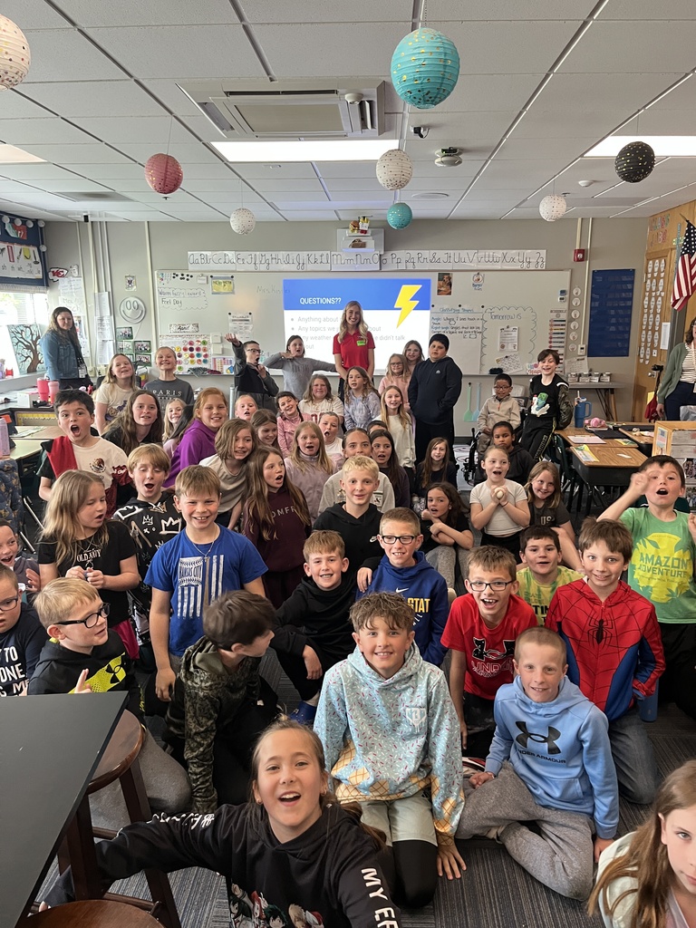 A large, energetic group of elementary students poses for a wide-angle class photo in a classroom with hanging paper lanterns. A blonde female guest speaker in a red polo shirt stands smiling at the center-back, positioned in front of a white board displaying a "QUESTIONS??" slide with a lightning bolt icon.