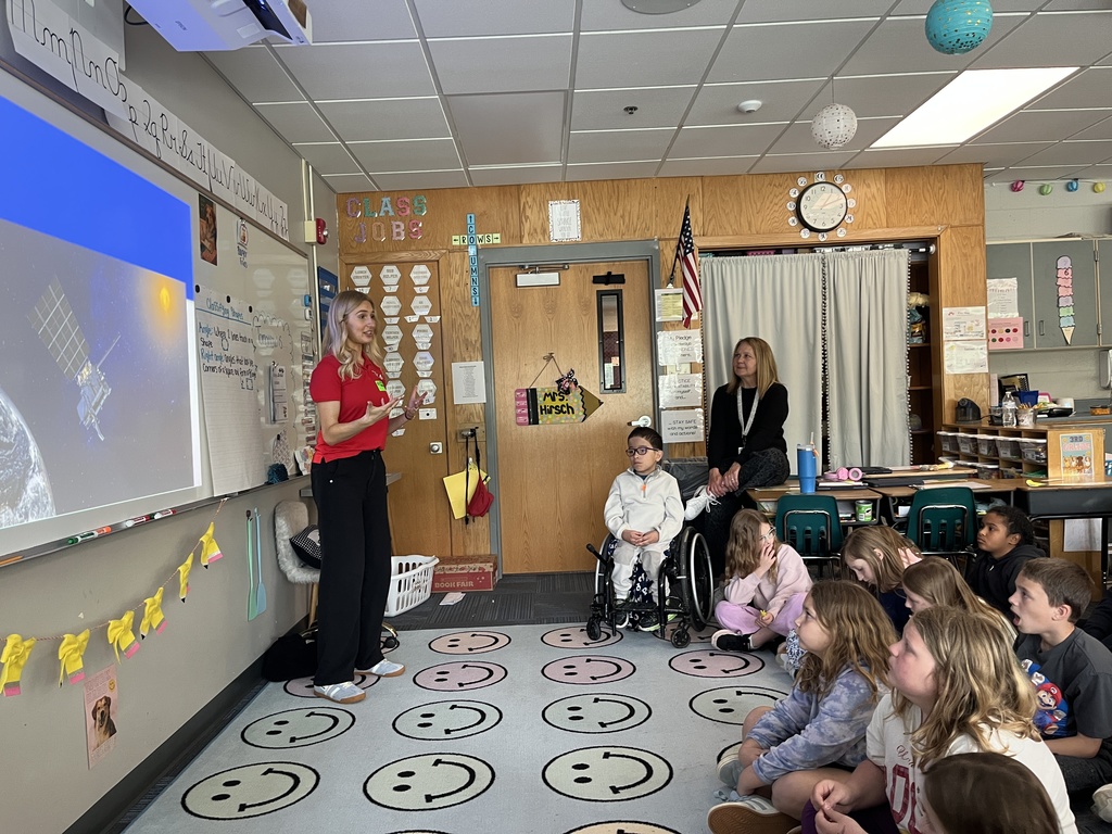 The guest speaker in the red polo stands by a projector screen showing a satellite in space, gesturing while speaking to the class. Students sit on a colorful rug patterned with smiley faces, and a teacher or aide observes from the background near a door labeled "Mrs. Hirsch."