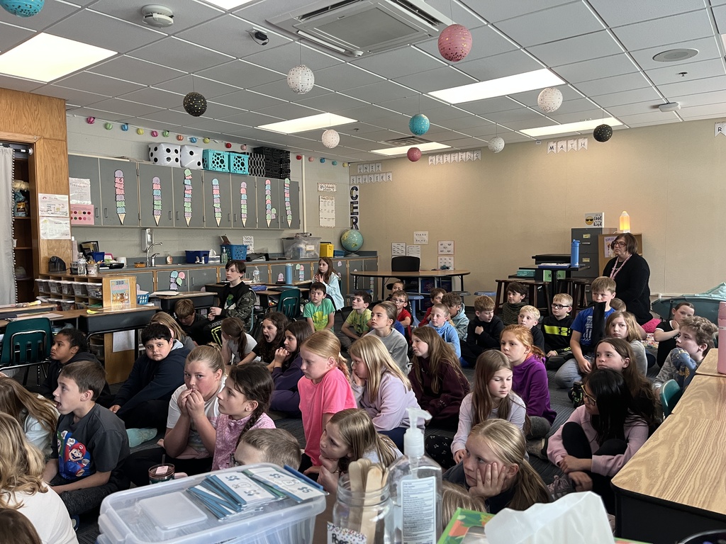 A side view of the classroom showing dozens of students sitting attentively on the floor, looking toward the speaker (off-camera). The room is decorated with "Class Jobs" and "Writing" displays, with educational posters and colorful storage bins lining the back wall.