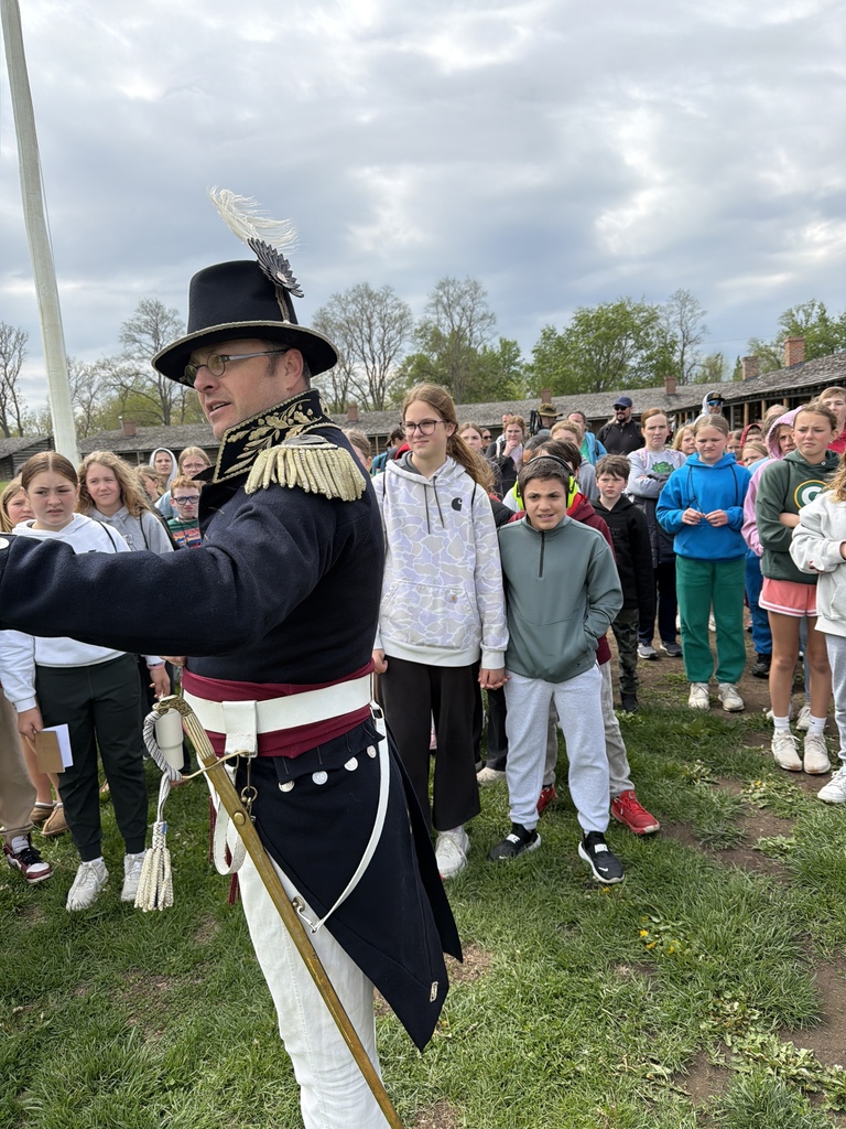 A man in a detailed historical military uniform and plumed hat speaking to a large group of students outdoors.