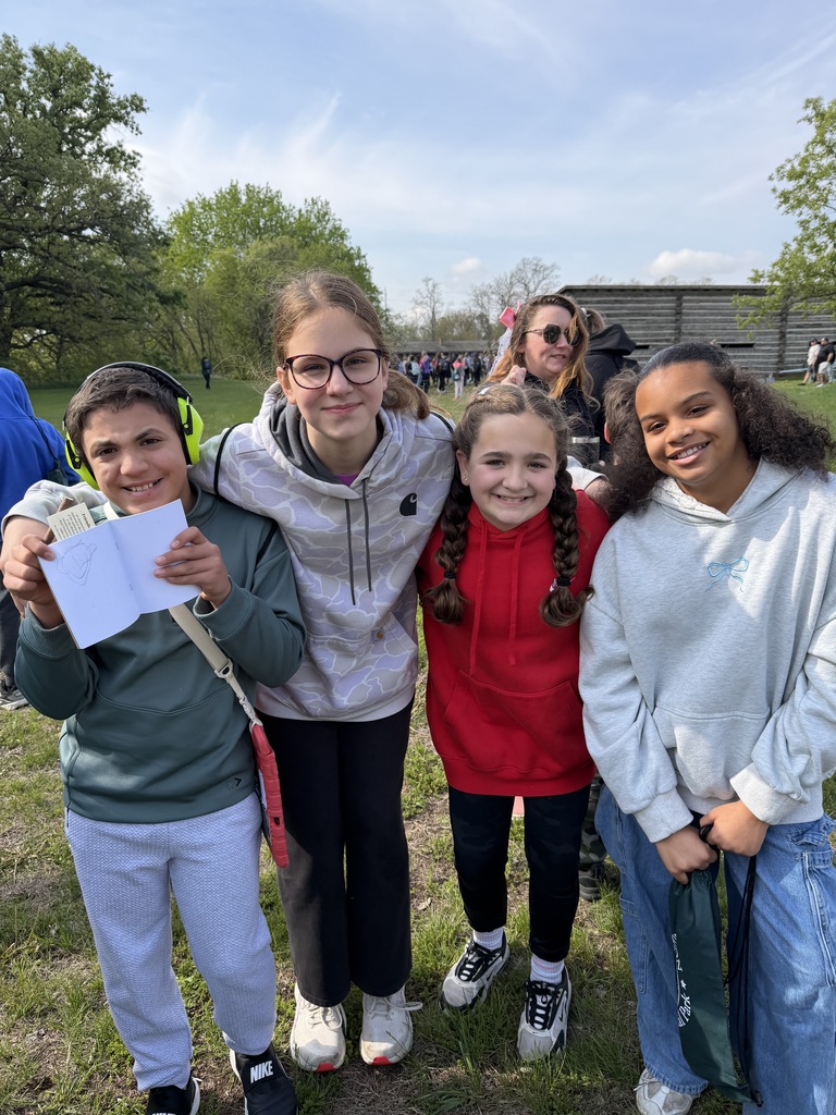 Four children huddling together and smiling outdoors; one boy holds up an open notebook showing a drawing.