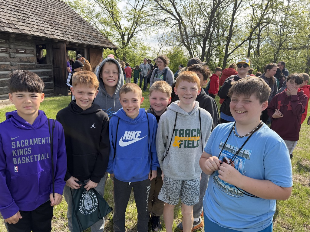 Seven young boys smiling for a group photo in front of a rustic log cabin during an outdoor field trip.