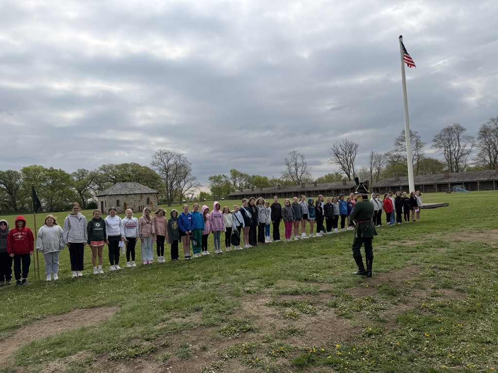A long line of students standing at attention in a field facing a man in a historical military uniform near a tall flagpole.