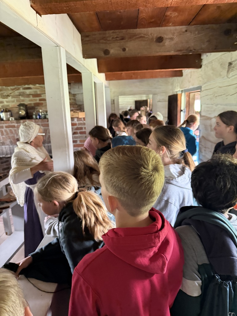 A crowd of students inside a historic log building listening to a woman dressed in period-accurate 19th-century clothing.