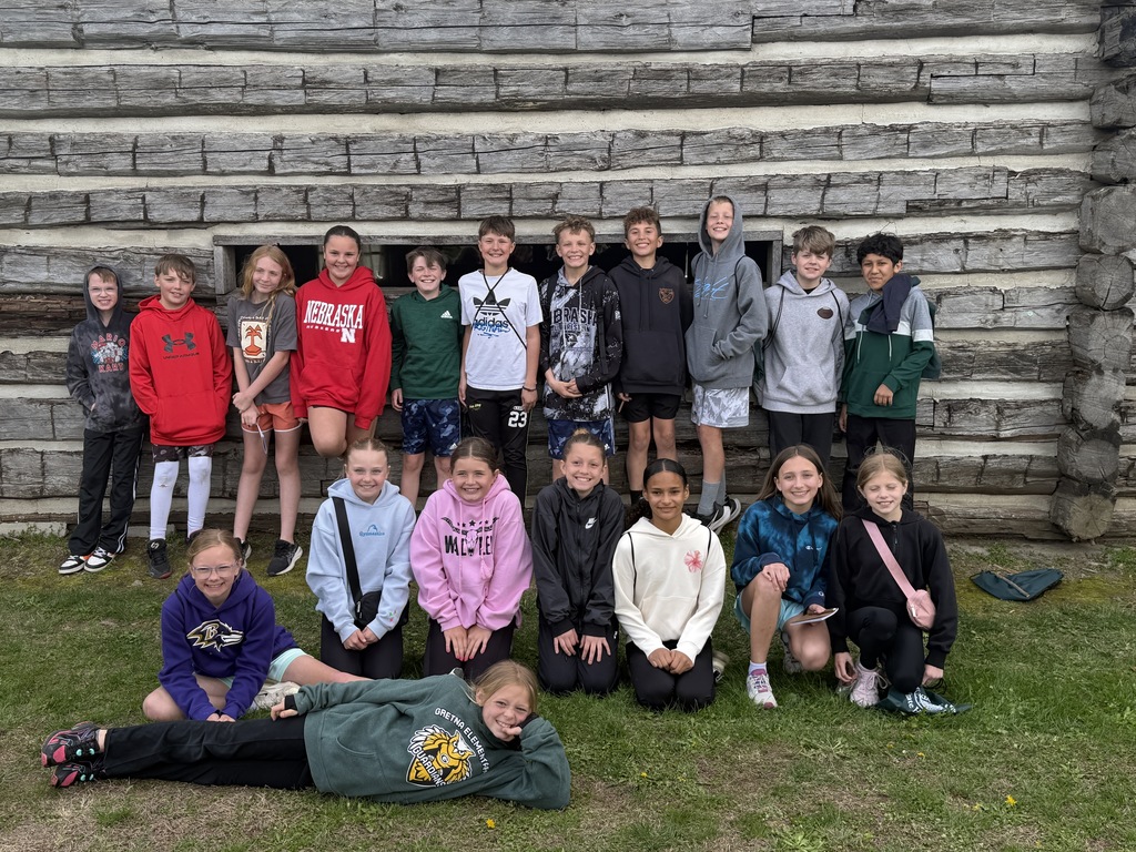 A group of students posing in front of a log building, with several girls kneeling in the front and one girl reclining on the grass.