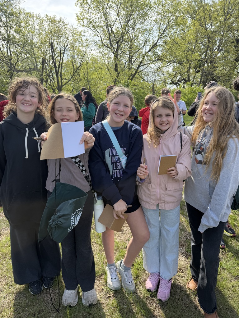 Five smiling girls standing together in a grassy field, some holding small notebooks and pens.