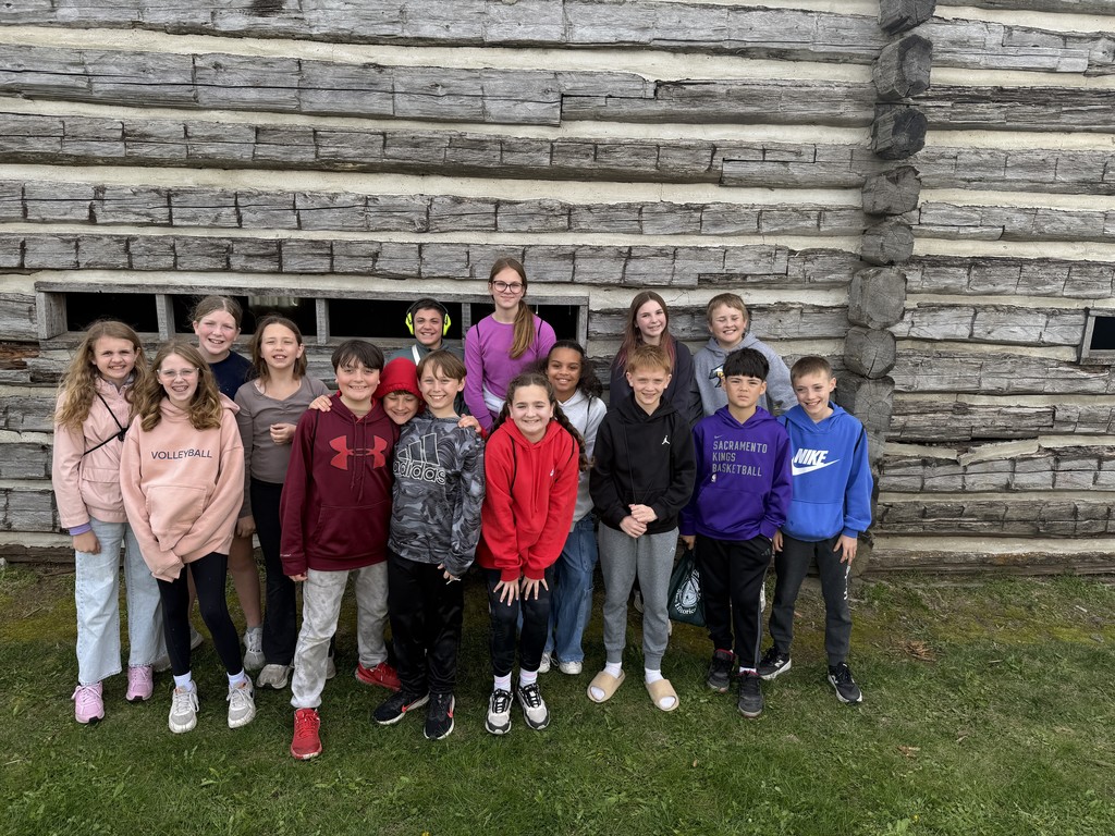 A group of 14 students posing together for a photo in front of a textured log wall.