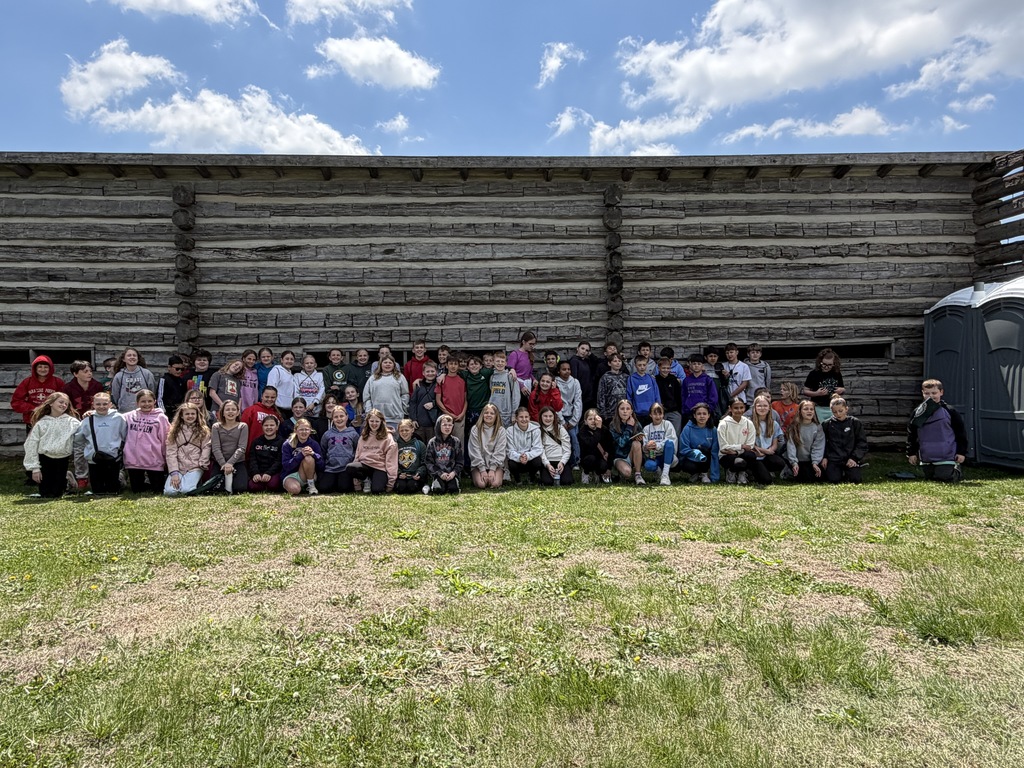 A wide group portrait of dozens of elementary-aged students posed in front of a long, historic log structure.