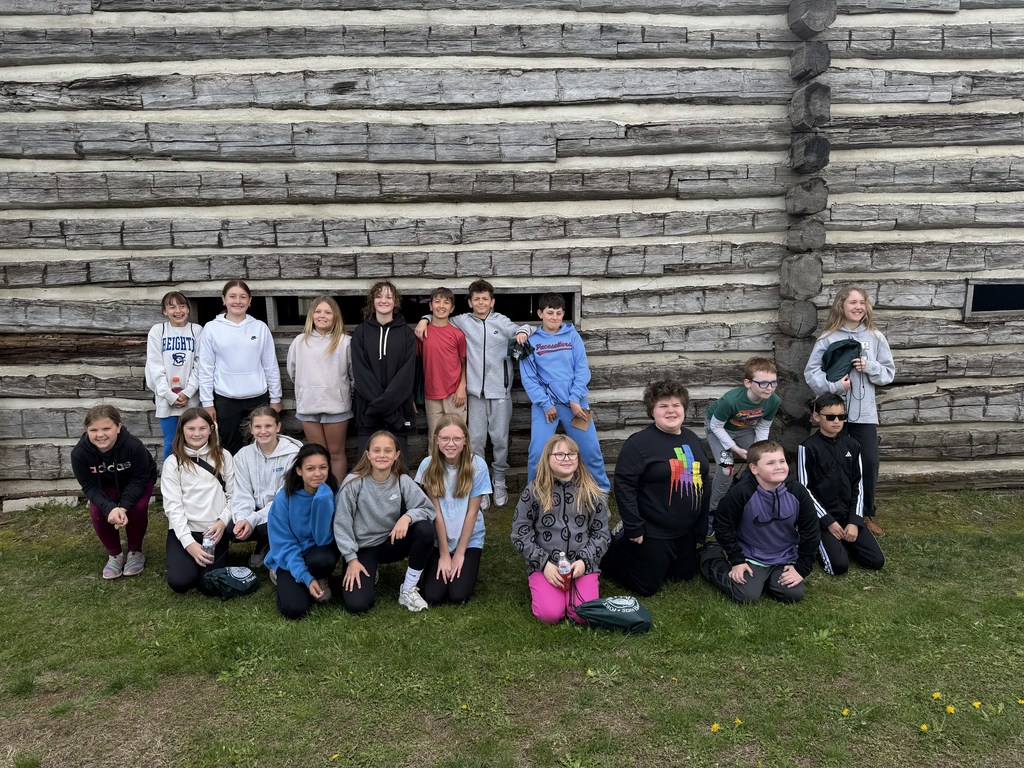 A group of 19 students in various colorful hoodies and jackets posing in two rows in front of a log building.