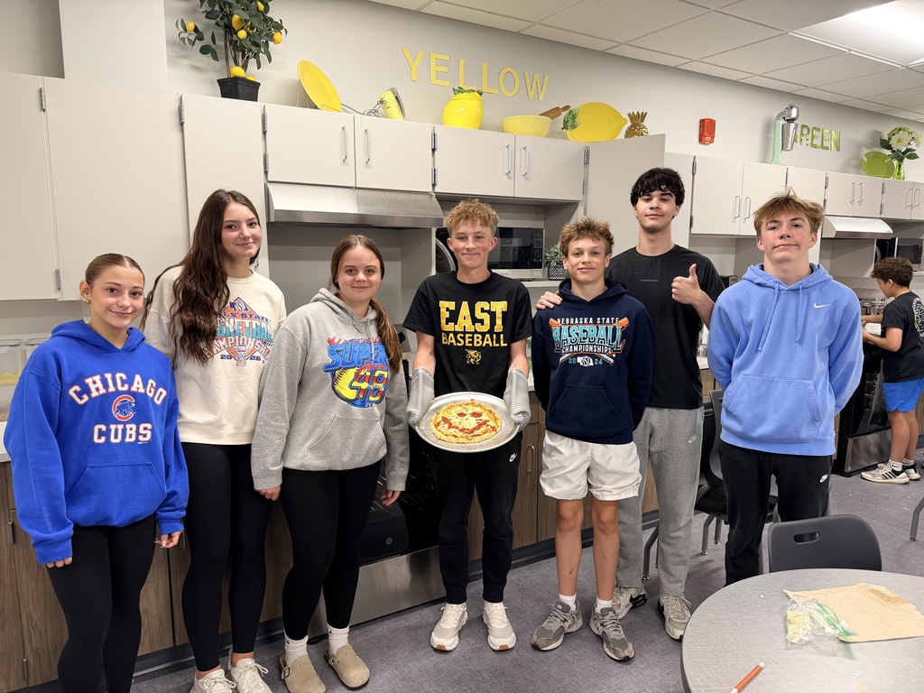 Students stand in a kitchen area and the one in the middle is holding a pizza with a smiley face made out of pepperonis. 