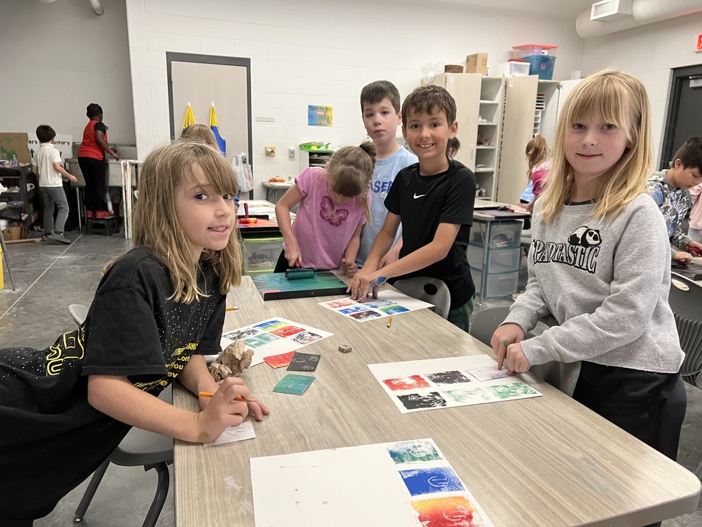 Three girls and two boys stand around their table pressing their prints onto their papers in art.  Papers have 3 rows of 3 squares.