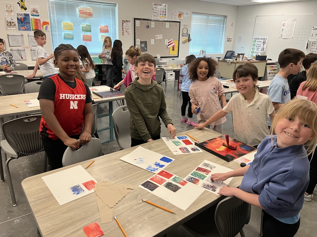 Three boys and two girls stand around their table pressing prints onto their paper.  Their papers are 3 rows of 3 squares.