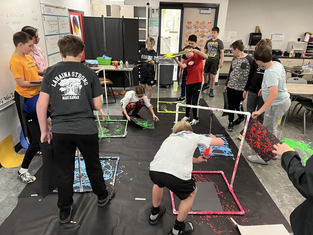 A group of fifth graders are observing students swinging paint bottles hanging from stands for pendulum painting.