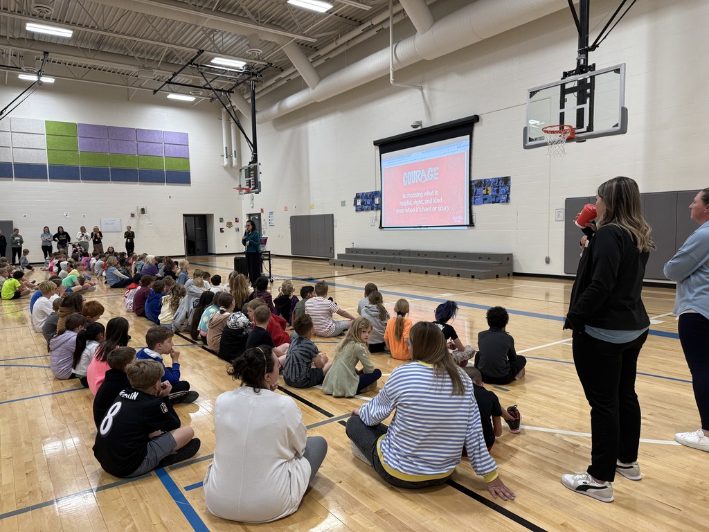 Rows of students sit in the gym looking at the principal and a sign showing Courage - it is choosing to do what is right, helpful or kind even when it's hard or scary.