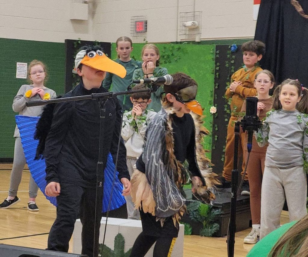 Two students in bird costumes with large orange beaks perform at a microphone during a school play.