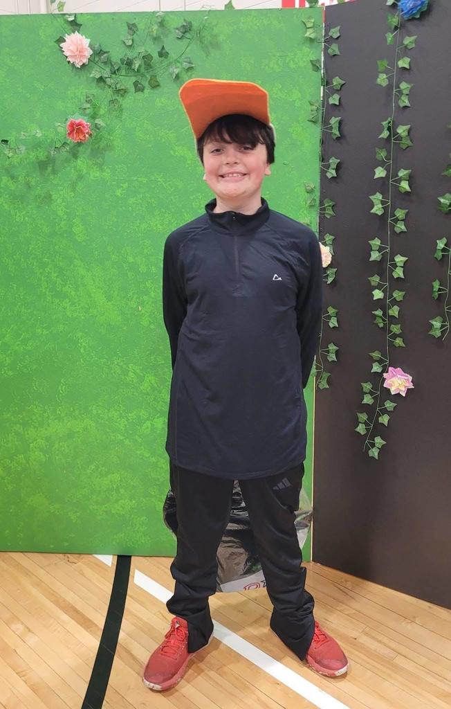 A boy in a black athletic pullover and an orange hat stands smiling in front of a green and black backdrop decorated with vines.