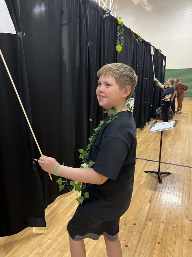 A boy wearing a black t-shirt and an ivy vine garland stands backstage near black curtains, holding a prop rod.