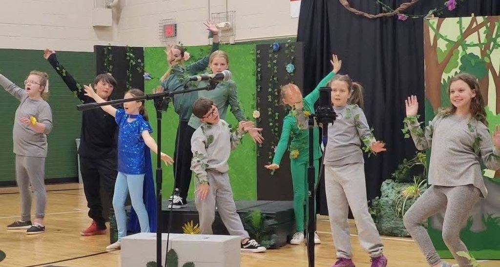 Several children in grey and green costumes with ivy accents perform a choreographed dance routine on stage.