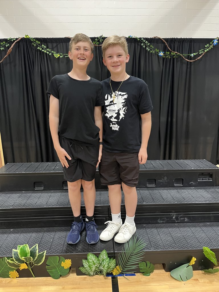 Two boys in black t-shirts stand on a stage riser decorated with jungle leaves; one wears a Jungle Book themed shirt.