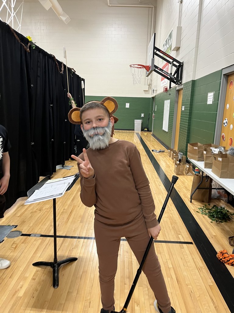 A boy in a brown thermal outfit, monkey ears, and a fake grey beard poses with a peace sign and a staff in a school gym.