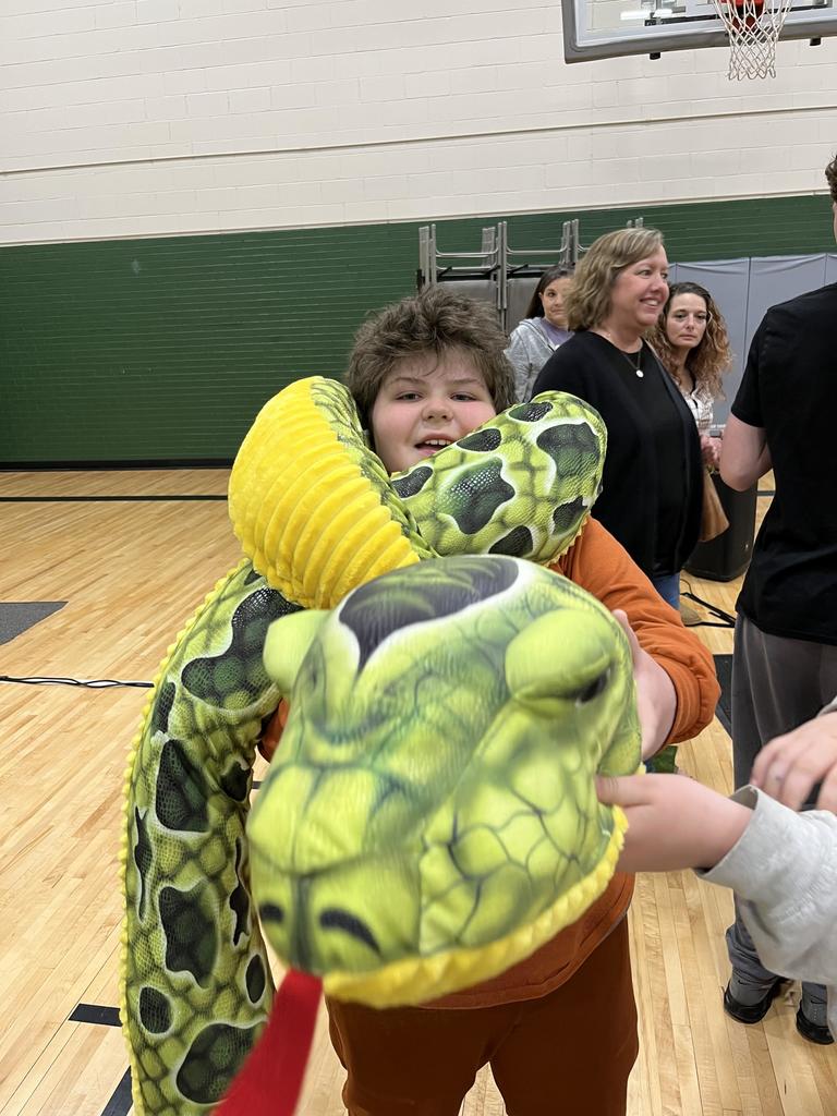 A student smiles while draped in a large, plush green and yellow toy snake in a gymnasium.