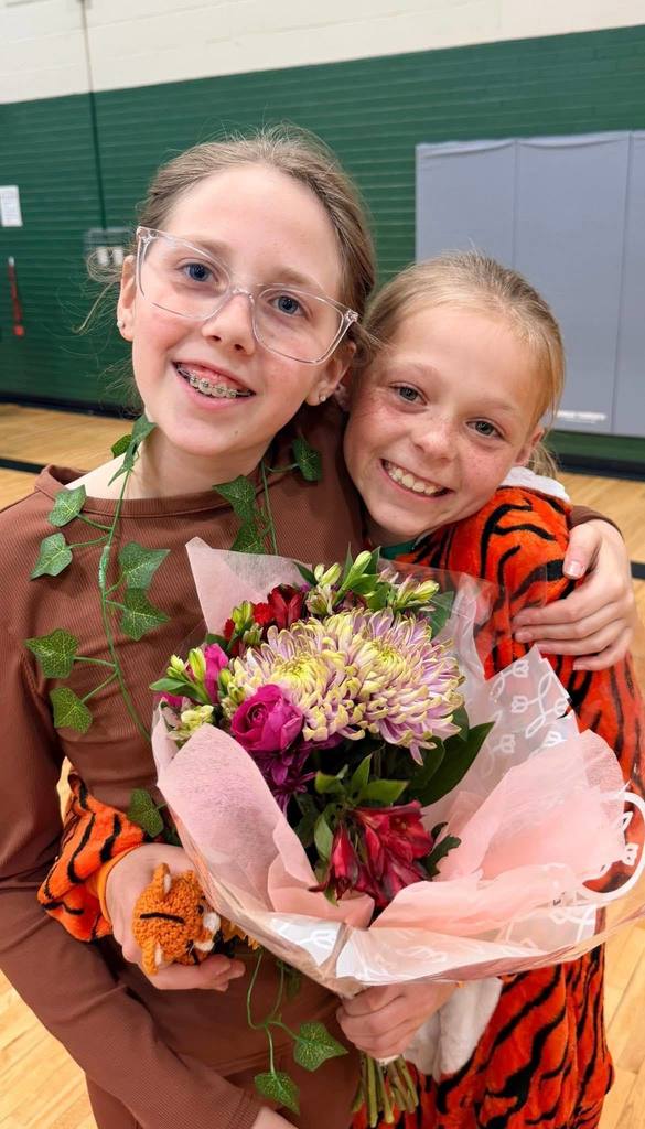 Two girls smile for a close-up photo; one wears a brown top with ivy and the other a tiger costume, while holding a bouquet of pink and purple flowers.