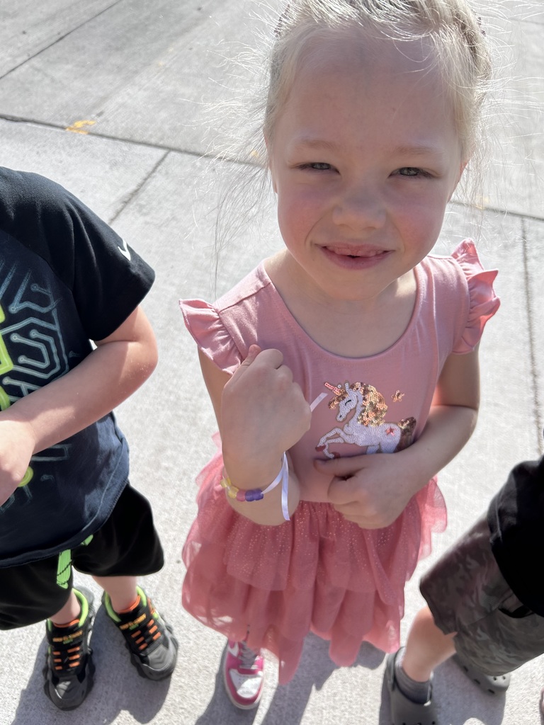 A girl in a pink unicorn dress with ruffled sleeves smiles at the camera while showing off her beaded bracelet.