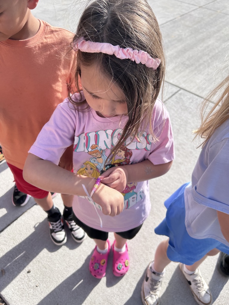 A young girl in a pink Disney Princess shirt and a pink headband looks down at her wrist as she adjusts a beaded bracelet.