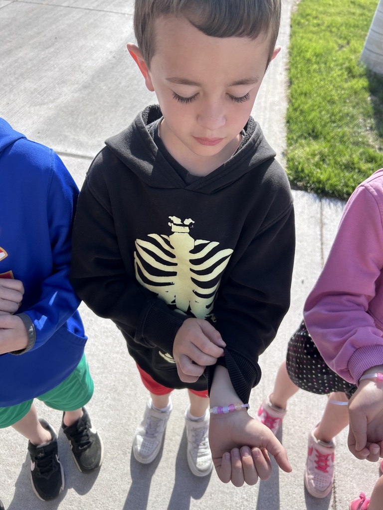 A young boy in a black skeleton-print hoodie looks down at a beaded bracelet on his wrist, standing on a sidewalk.