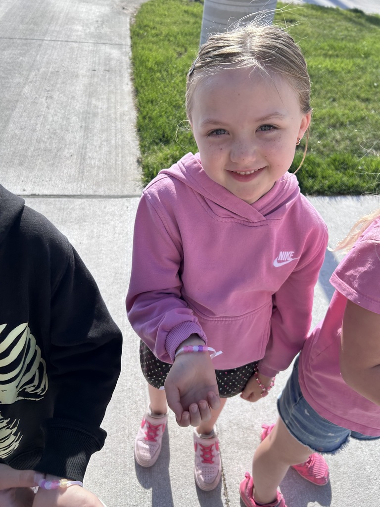 A smiling girl in a pink Nike hoodie stands outdoors, holding out her wrist to show a handmade bracelet with pink and purple beads.