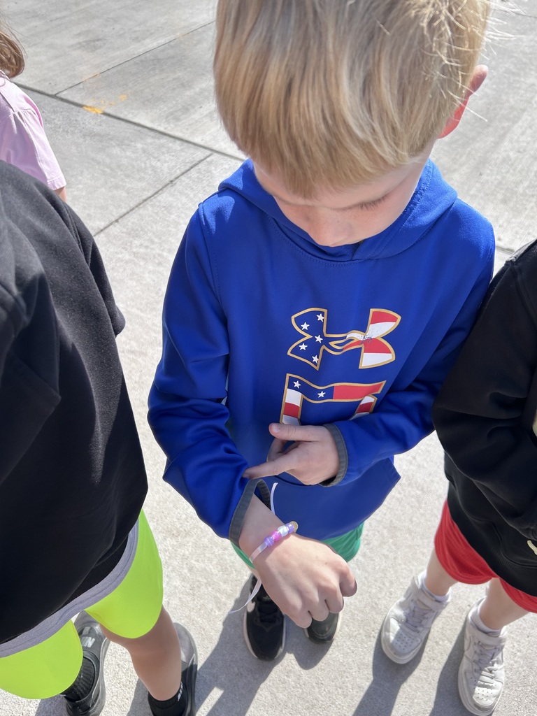 A boy wearing a blue Under Armour hoodie with a patriotic logo points to a pink and purple beaded bracelet on his wrist.
