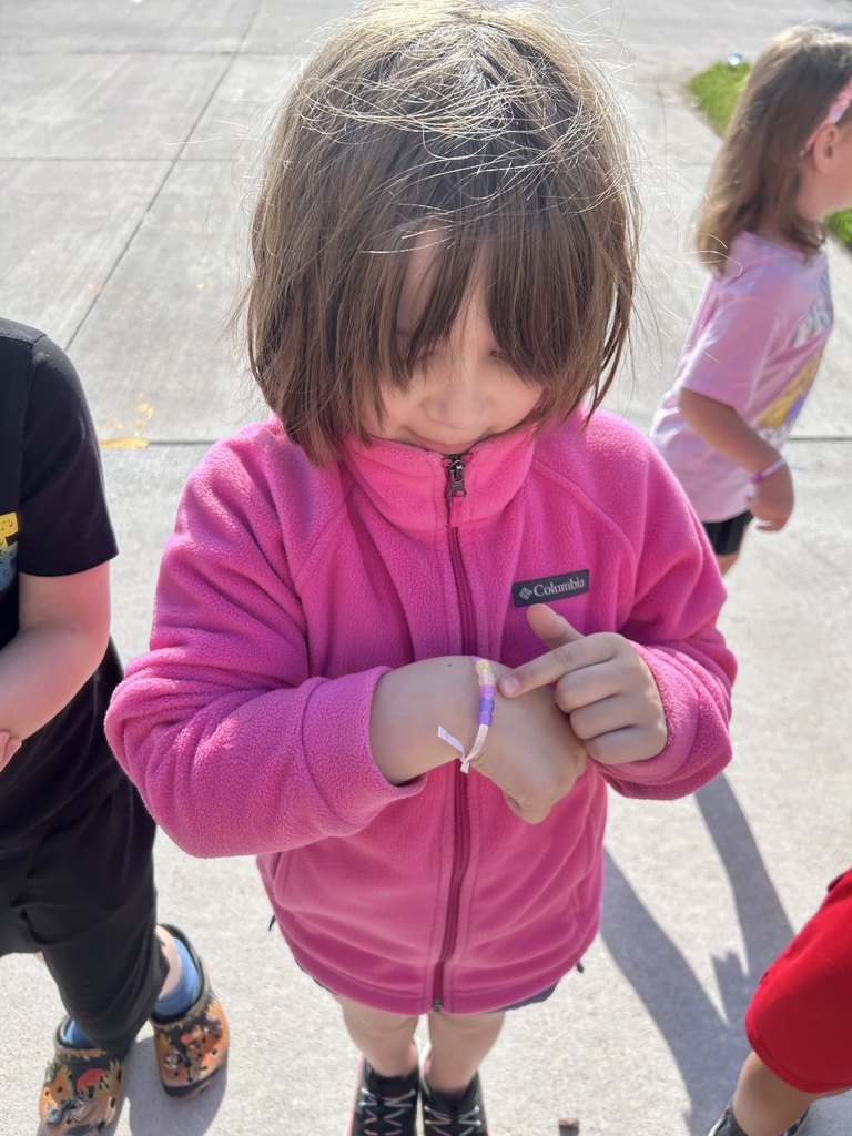 A girl in a bright pink Columbia fleece jacket looks down and points at her colorful beaded bracelet.