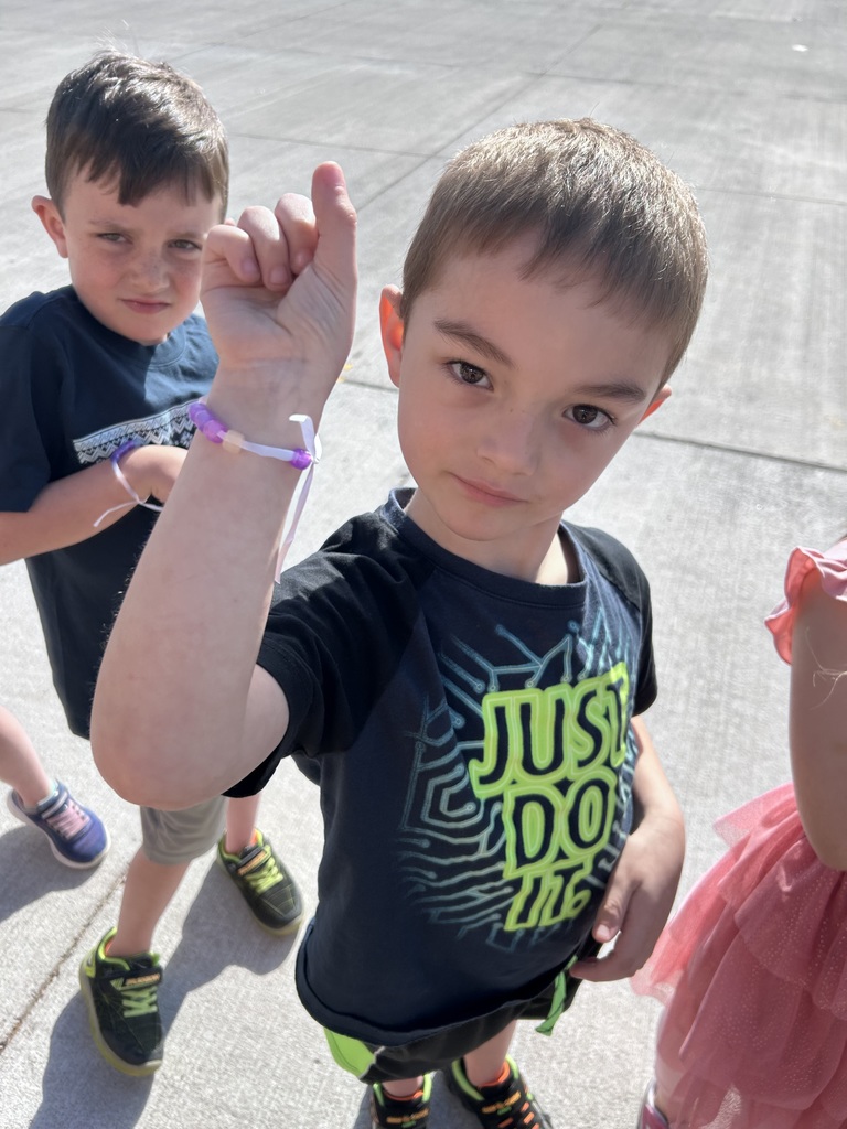 A boy in a black "Just Do It" t-shirt raises his arm to display a purple and white beaded bracelet; another child stands in the background.