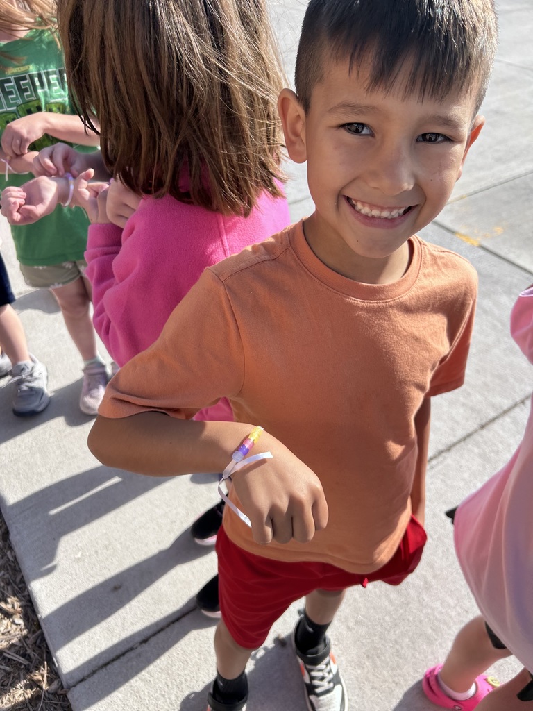 A smiling boy in an orange t-shirt displays a bracelet made of yellow, purple, and pink beads on his wrist.