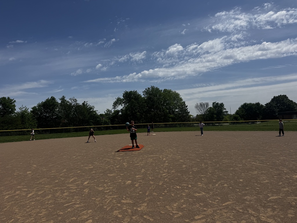 Wide shot of a youth kickball or baseball game in progress on a dirt field under a bright blue, wispy sky.