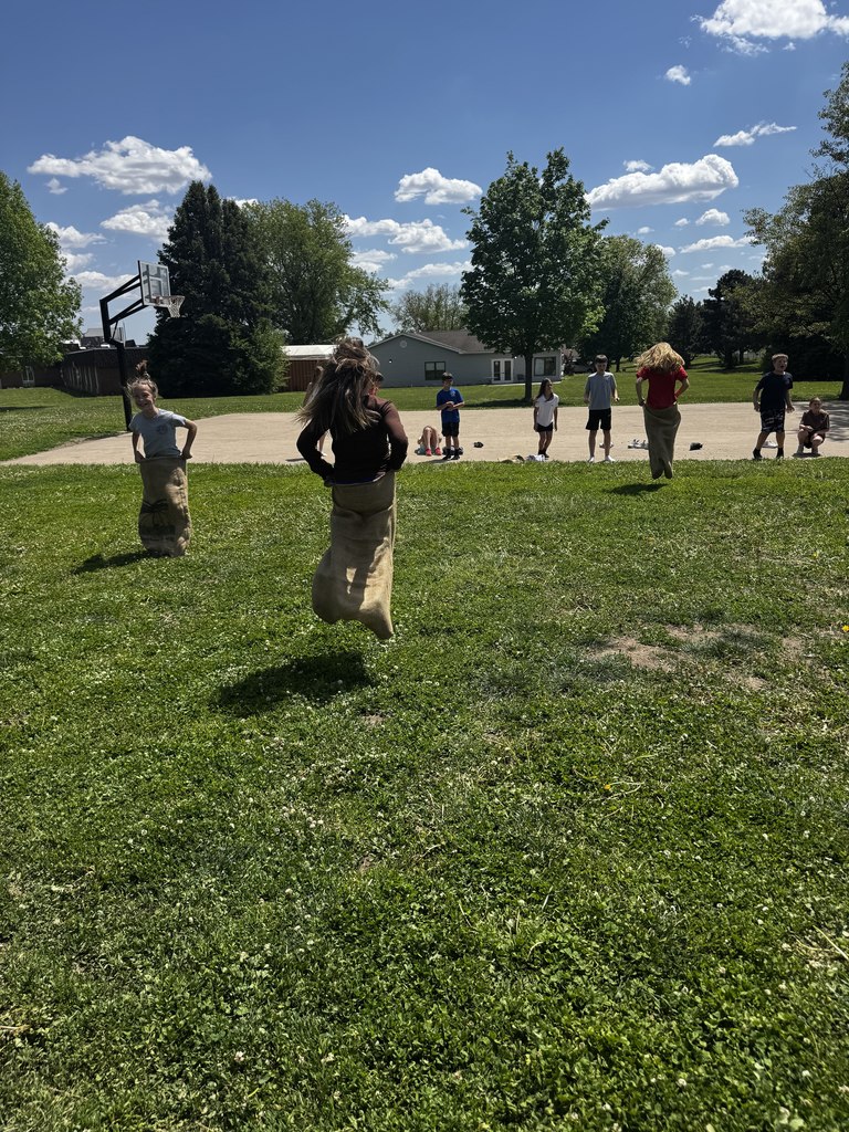 Three children in burlap sacks competing in a race on a bright, grassy field with a basketball hoop in the background.