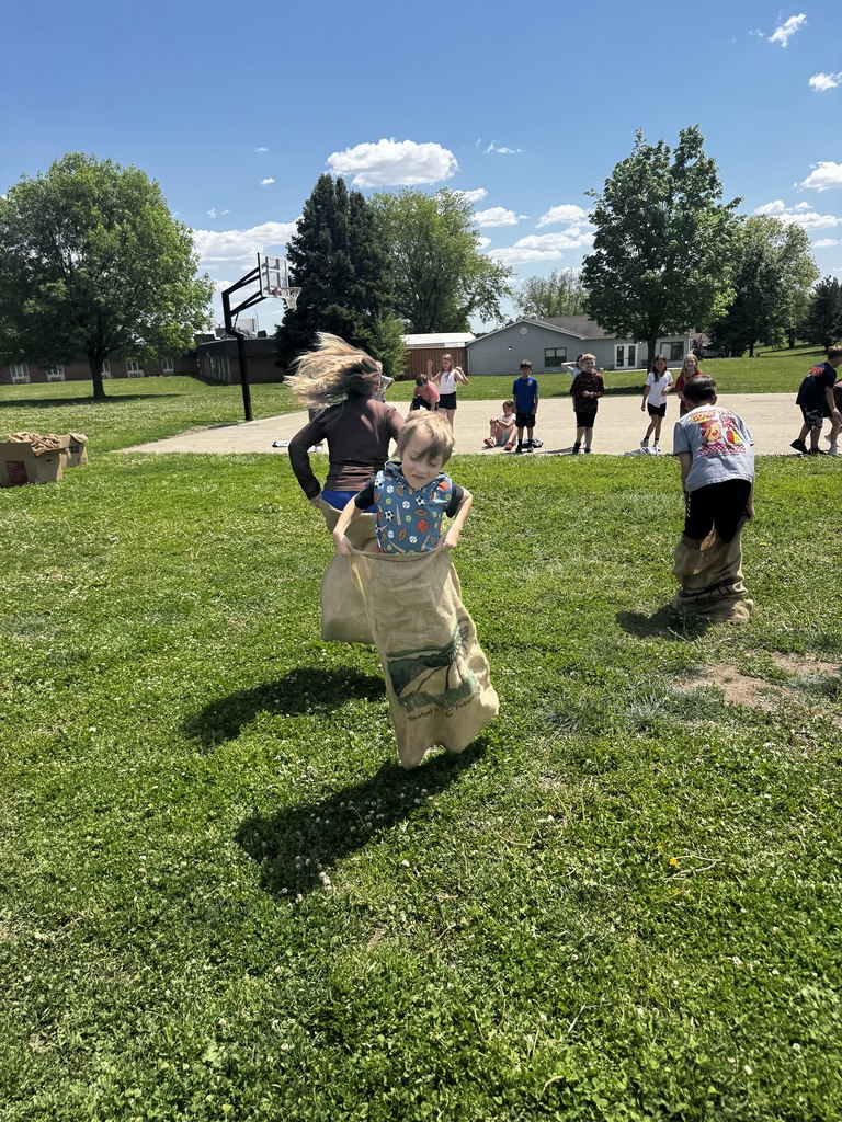 Three children in burlap sacks competing in a race on a bright, grassy field with a basketball hoop in the background.