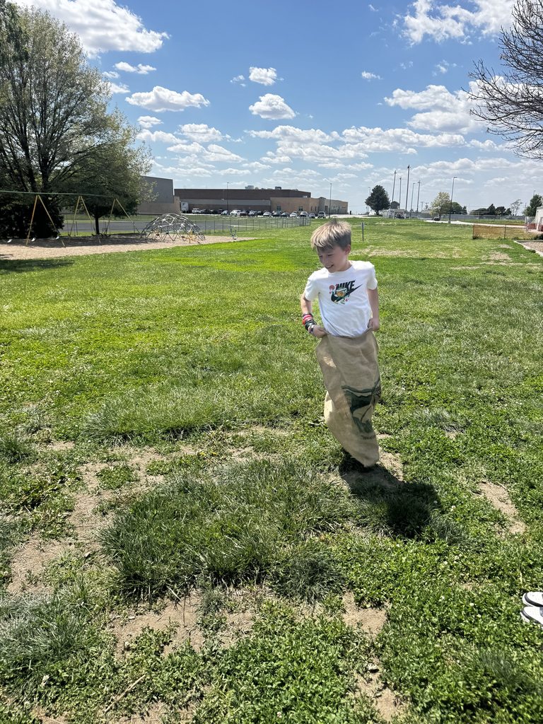 A smiling boy in a white Nike t-shirt participating in a sack race on a large green lawn near a school playground.