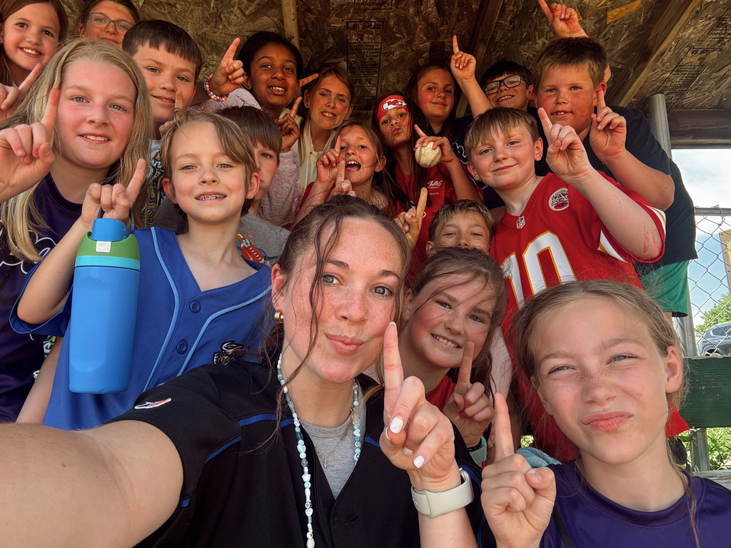 A large group of children and a woman taking a crowded, celebratory selfie in a dugout, everyone holding up one finger.