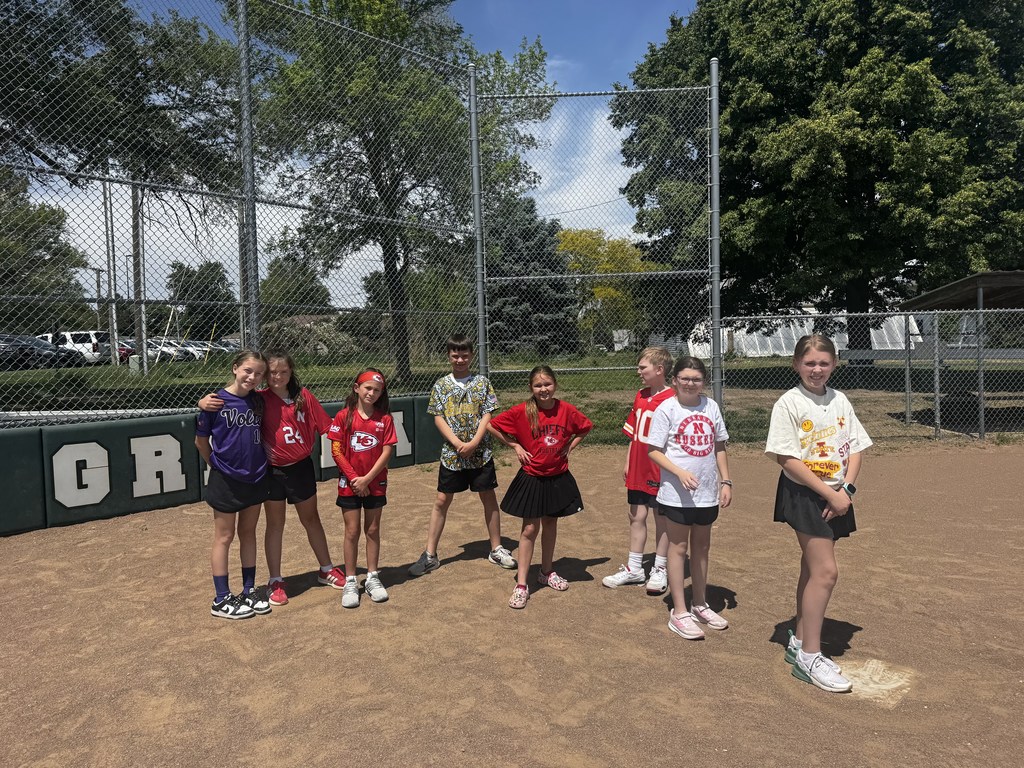 A group of eight children in colorful sports jerseys posing for a group photo on a dirt baseball infield.