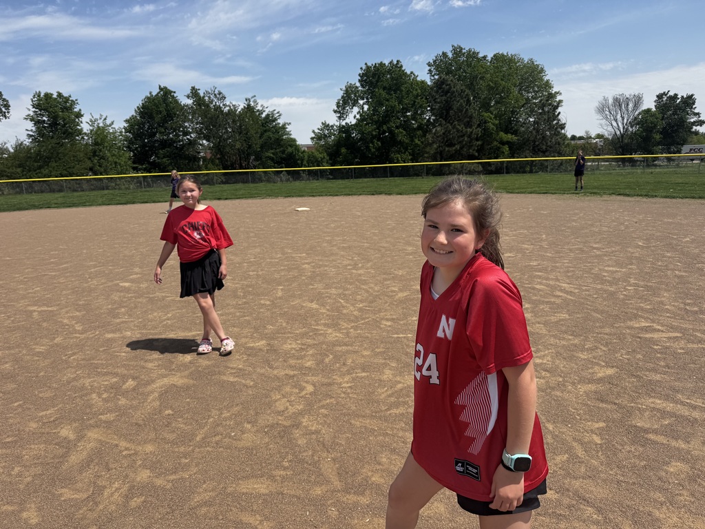 A young girl in a red "Husker" jersey smiling on a dirt field with a teammate standing in the background.