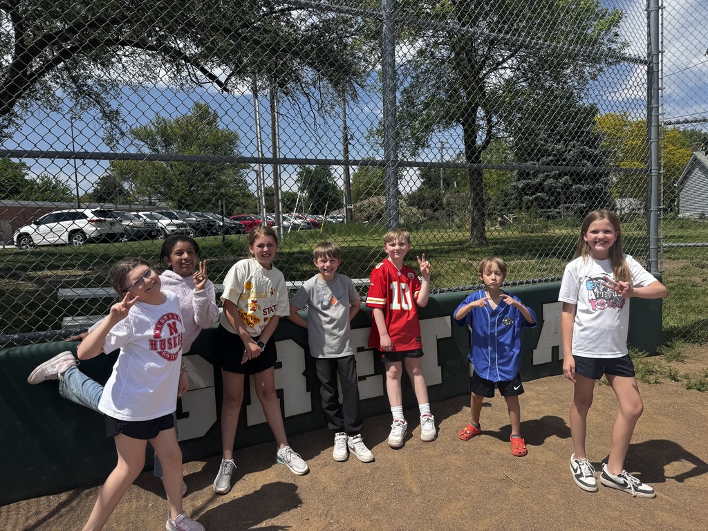 Seven children posing and making peace signs against a green outfield fence with "GRETNA" visible on the padding.