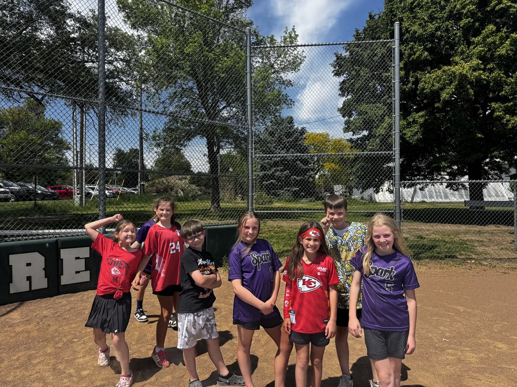 Seven children in purple and red team jerseys posing together on a dirt sports field in front of a chain-link fence.