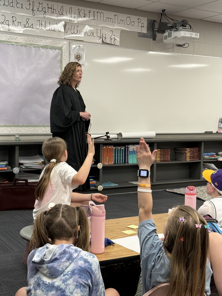 A woman in a black judicial robe stands at the front of a classroom, speaking to a group of seated students who are raising their hands. A gavel and law books are visible on the shelf behind her.