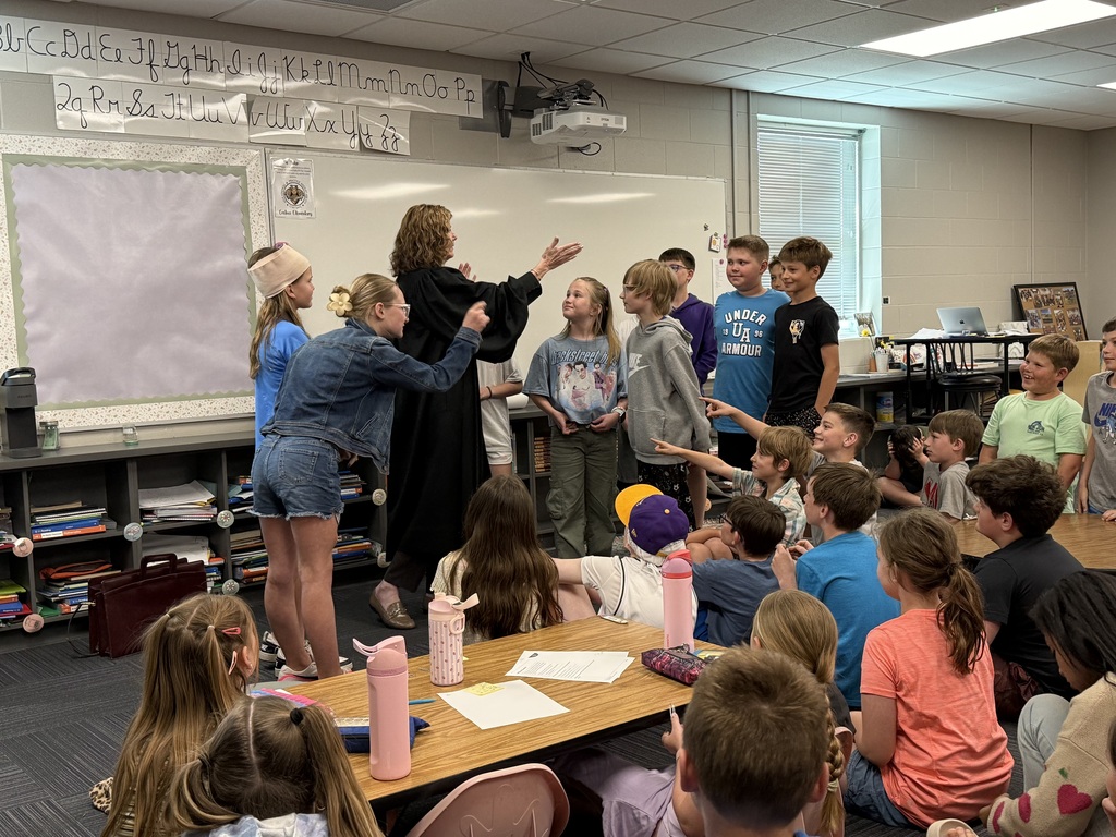 A woman in a judicial robe facilitates a mock trial in a classroom, with students standing and gesturing toward one another while their classmates watch from the floor and desks.