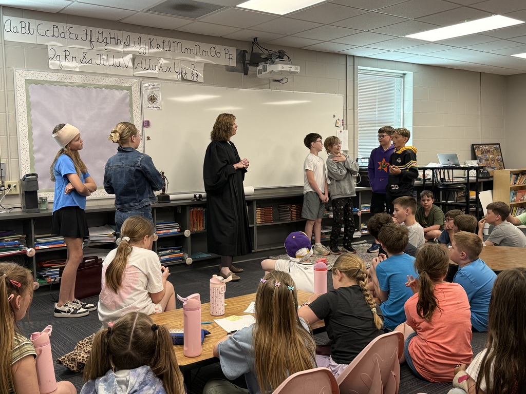 A woman in a judicial robe stands before a group of elementary students in a classroom, where several children are standing in a line as if participating in a mock trial or presentation.