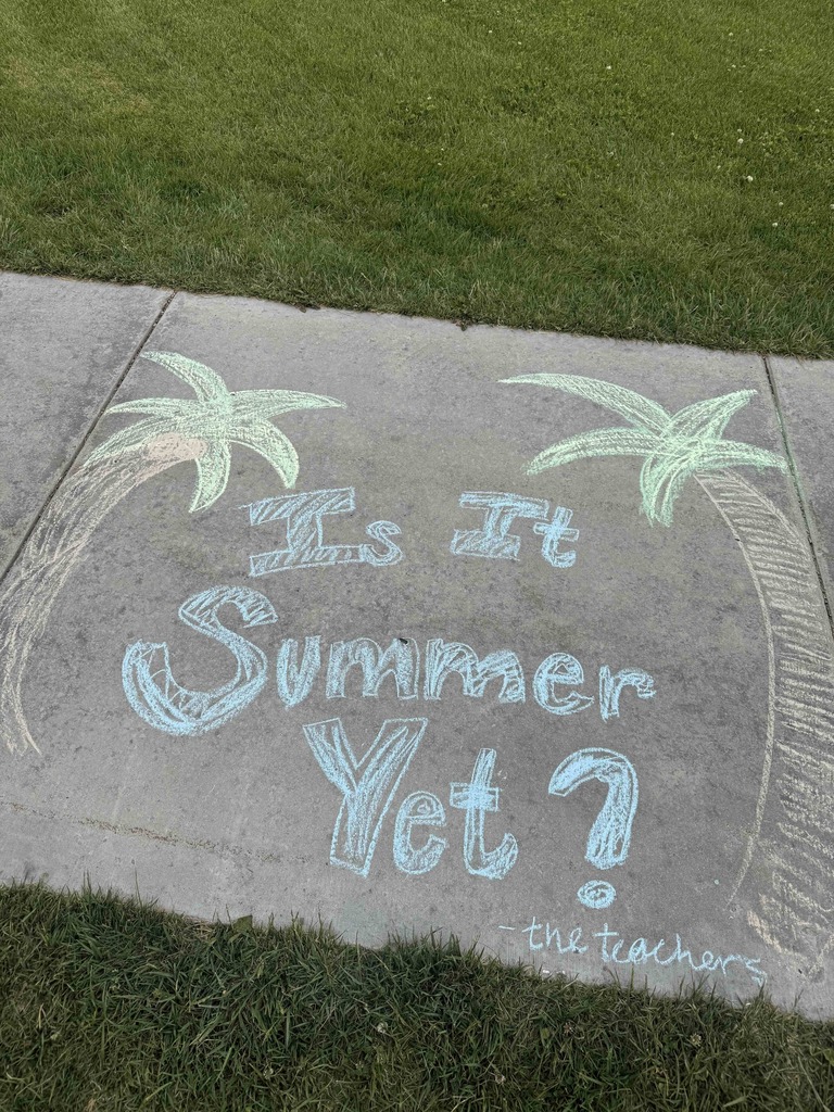 A close-up of a completed chalk drawing on a sidewalk square. The message "Is It Summer Yet?" is written in large, bubbly blue letters, framed by two hand-drawn palm trees. In the bottom right corner, it is humorously signed "-the teachers" in smaller white script.
