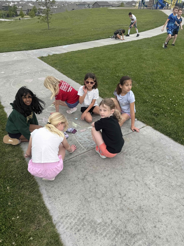 A group of five young girls sits in a circle on a sidewalk path, collaborating on a chalk drawing. They are surrounded by green grass, and in the background, other students are visible playing and drawing further down the path near a school playground.