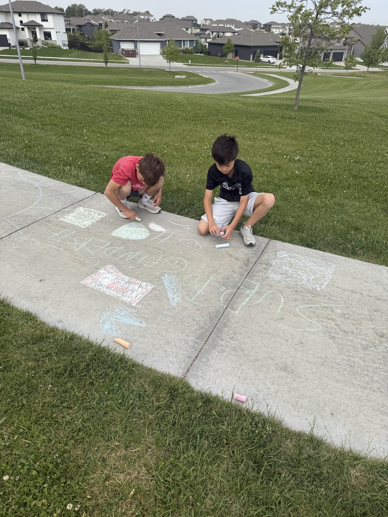 Two boys kneel on a sidewalk, adding details to a large collection of chalk drawings. One boy in a red shirt draws a square, while the other in a black shirt works on a nearby design. The sidewalk is covered in various shapes and faint words, with a neighborhood and rolling green hills visible behind them.